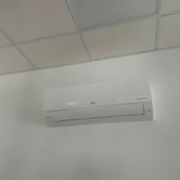 Biała klimatyzacja LG Dual Inverter zamontowana na białej ścianie w pomieszczeniu z kasetonowym sufitem. Widoczny nawiewnik sufitowy.