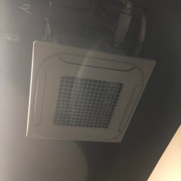 MR INSTAL - Kasetonowa jednostka klimatyzacji sufitowej z widocznymi przewodami, zamontowana w ciemnym pomieszczeniu. Biała kratka nawiewu w kwadratowej obudowie.