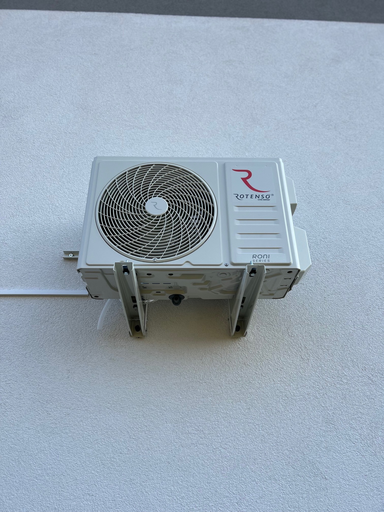 Zamontowana jednostka zewnętrzna klimatyzatora Rotenso Roni Series na białej ścianie z widocznymi elementami montażowymi i przewodem.