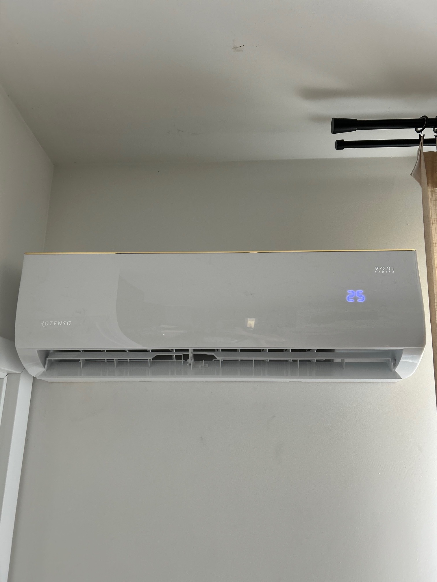 Zamontowany klimatyzator ROTENSO Roni Series na ścianie, wyświetlający temperaturę 25°C. Widoczne elementy instalacji i zasłona w tle.