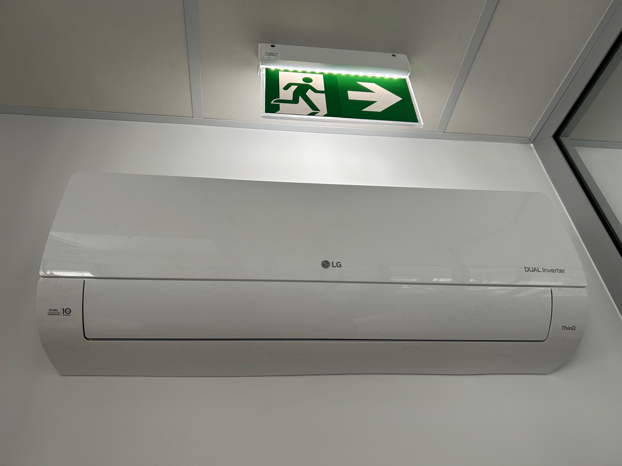 Biała klimatyzacja LG Dual Inverter zamontowana na ścianie, podświetlony znak ewakuacyjny nad urządzeniem. Widoczny fragment okna w prawym górnym rogu.