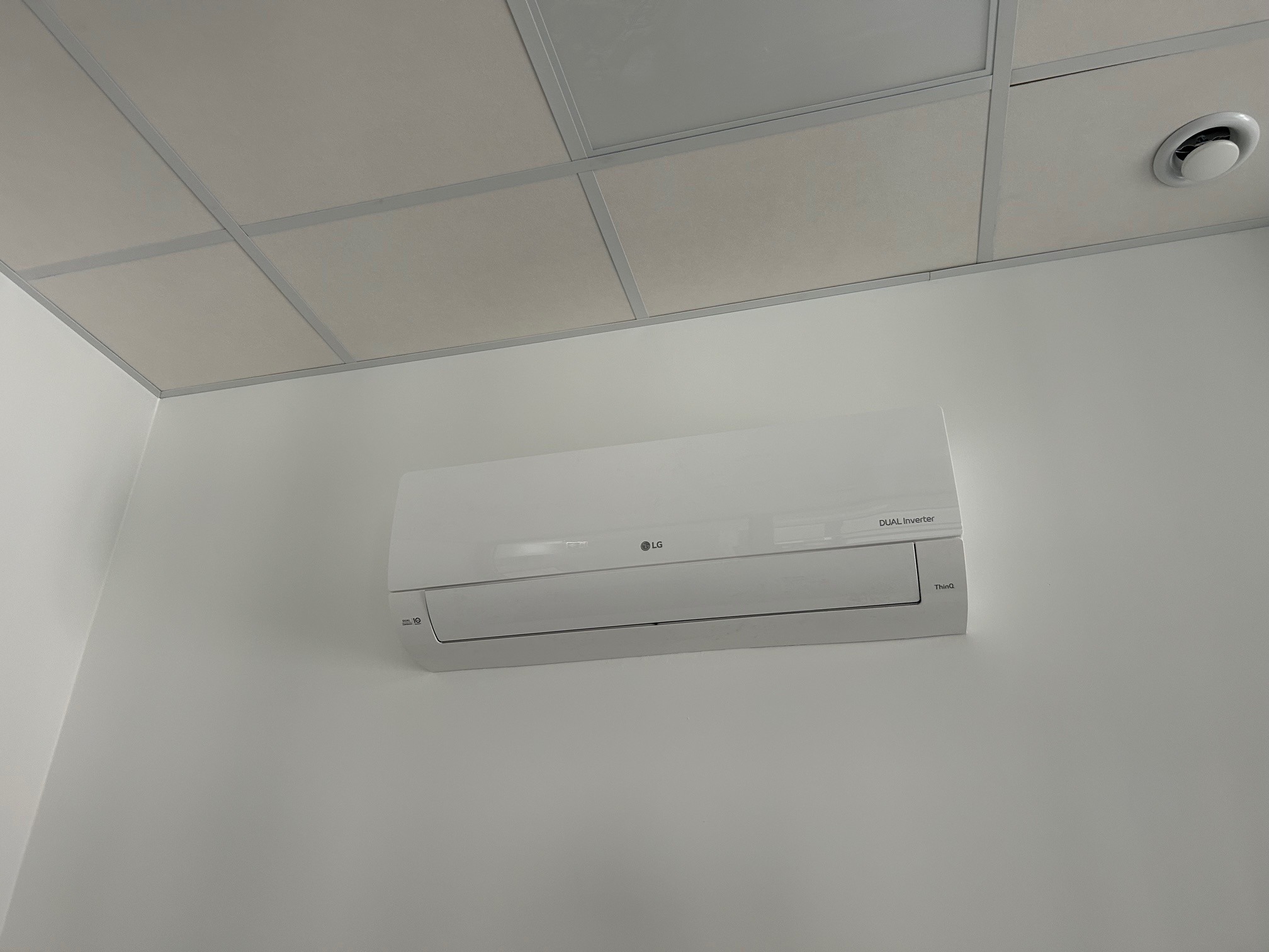 Biała klimatyzacja LG Dual Inverter zamontowana na białej ścianie w pomieszczeniu z kasetonowym sufitem. Widoczny nawiewnik sufitowy.