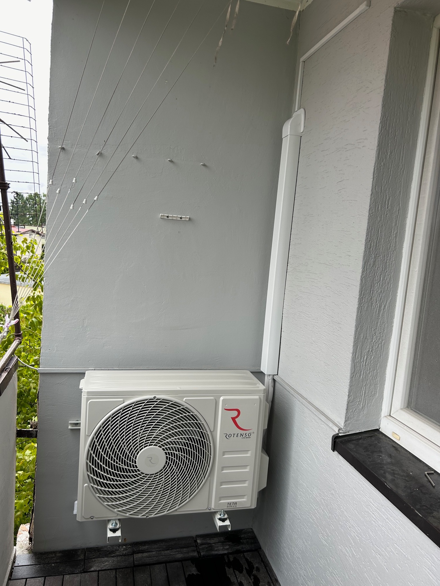 Biała jednostka zewnętrzna klimatyzacji Rotenso, zamontowana na szarym balkonie z drewnianą podłogą. Na ścianie widoczne linki do suszenia prania i okno.