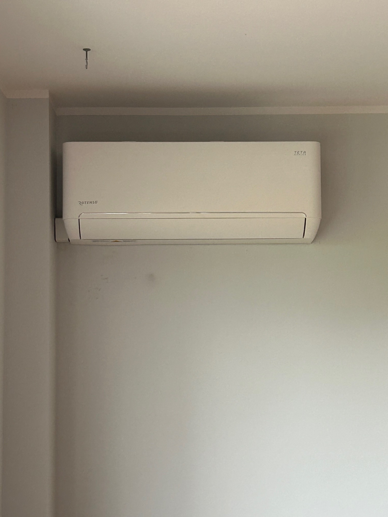 Biały klimatyzator Rotenso Iria zamontowany na jasnej ścianie w pomieszczeniu. Widoczny hak w suficie. Minimalistyczny design.