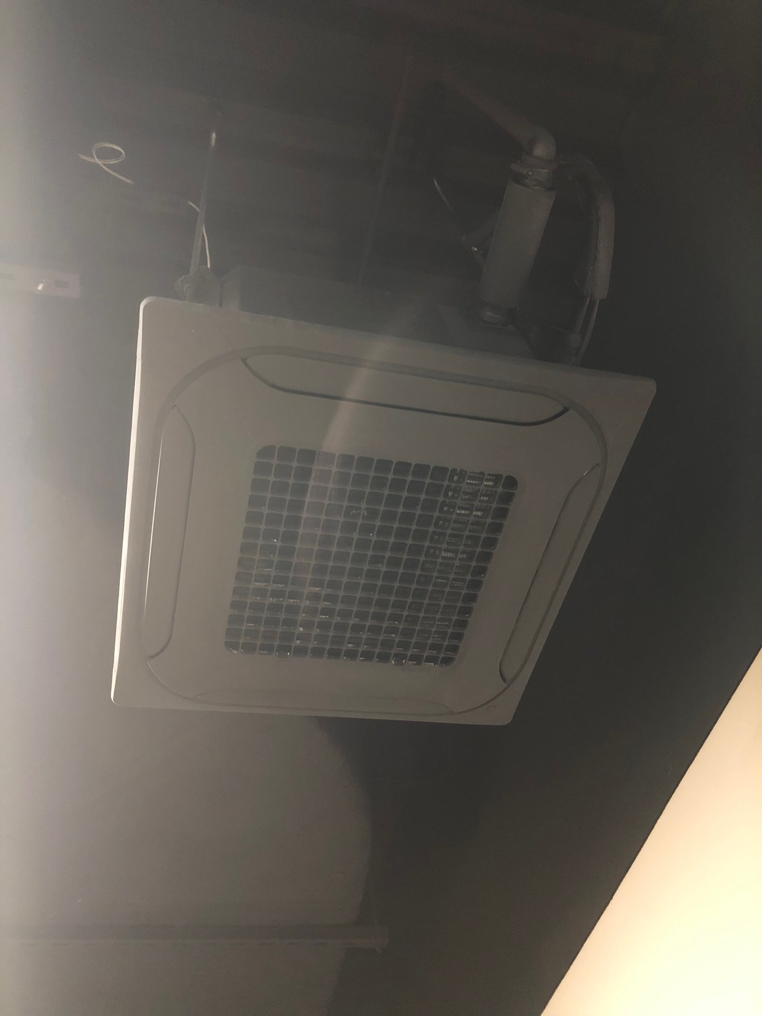 Kasetonowa jednostka klimatyzacji sufitowej z widocznymi przewodami, zamontowana w ciemnym pomieszczeniu. Biała kratka nawiewu w kwadratowej obudowie.