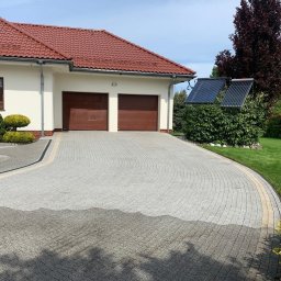 ABMax Alan Białecki - Świeżo umyta, jasnoszara kostka brukowa ułożona w łukowaty wzór przed domem z czerwoną dachówką i dwoma brązowymi bramami garażowymi, widoczne mokre plamy po myciu.