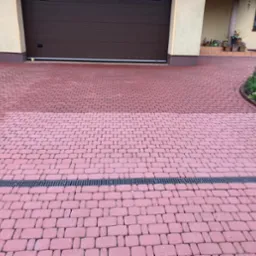 Częściowo wyczyszczona, czerwona kostka brukowa na podjeździe przed garażem, z widoczną różnicą w kolorze między czystą a brudną powierzchnią, oddzielona czarną kratką odpływową.