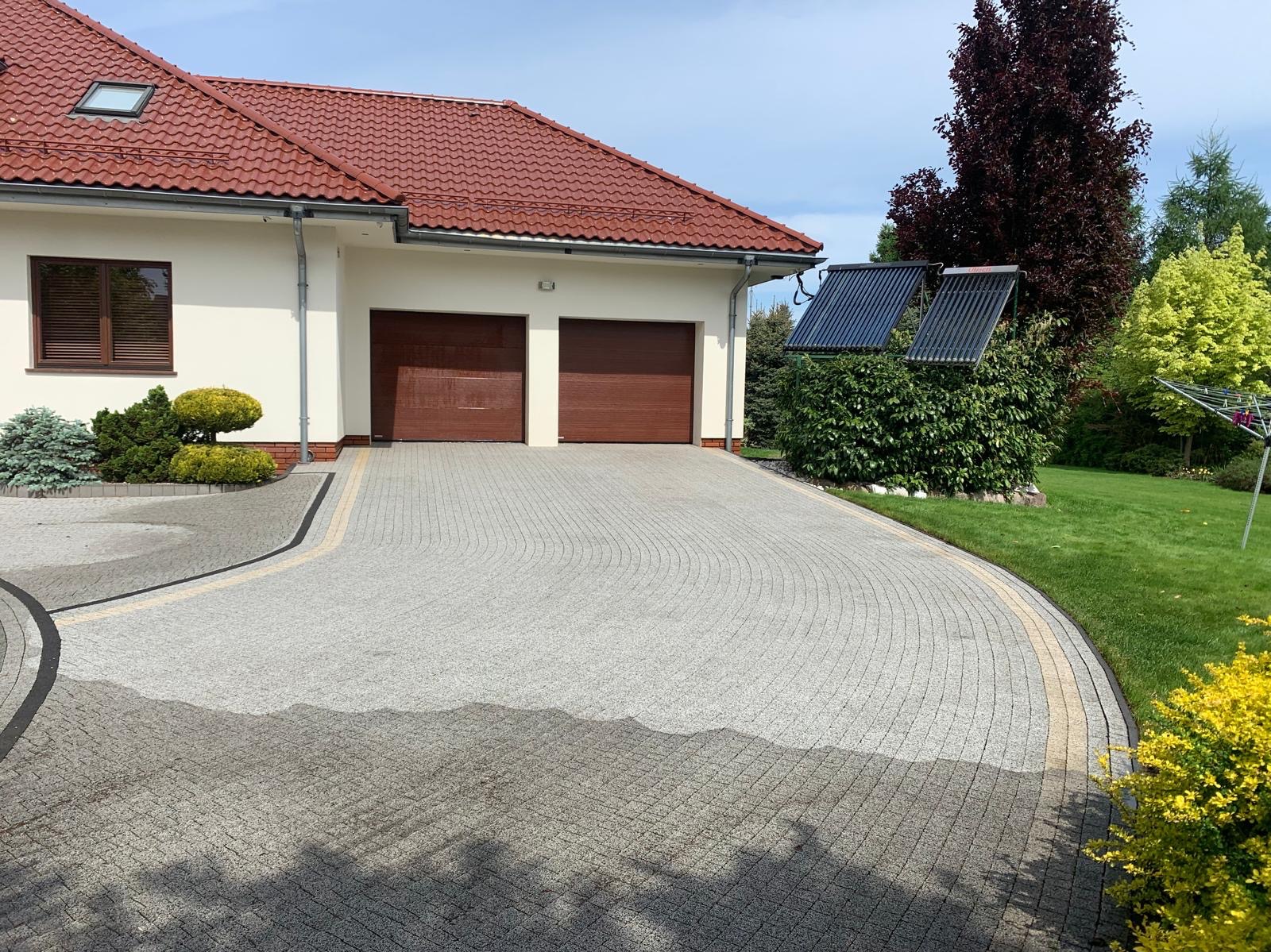 Świeżo umyta, jasnoszara kostka brukowa ułożona w łukowaty wzór przed domem z czerwoną dachówką i dwoma brązowymi bramami garażowymi, widoczne mokre plamy po myciu.