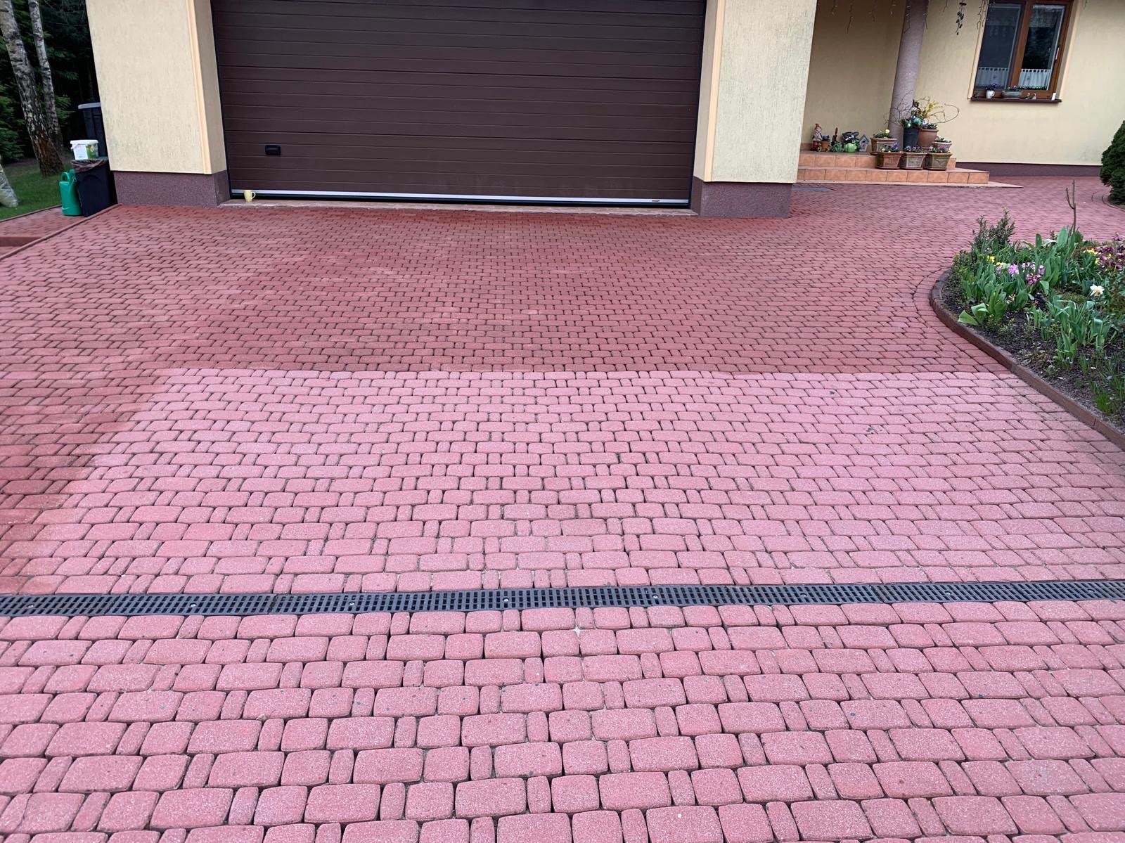 Częściowo wyczyszczona, czerwona kostka brukowa na podjeździe przed garażem, z widoczną różnicą w kolorze między czystą a brudną powierzchnią, oddzielona czarną kratką odpływową.