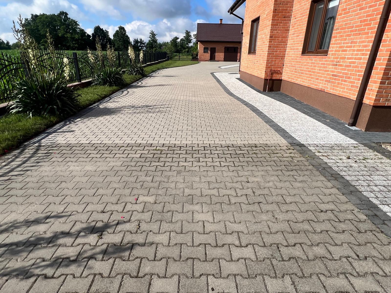 Szeroki kadr na szarą kostkę brukową prowadzącą do domu z czerwonej cegły z brązowymi oknami, widoczne lekkie zabrudzenia na kostce, z boku metalowe ogrodzenie z roślinami.