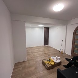 VinyllTech - Pokoje po remoncie z nową podłogą z paneli, widoczny fragment sofy z osobą, karton z narzędziami i akcesoriami malarskimi, oraz ozdobna ściana z pionowych listew.