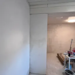 Wnętrze w trakcie prac wykończeniowych, widoczne ściany po nałożeniu gładzi, sufit z drewnianą boazerią, podłoga przygotowana do dalszych prac, dwa wiadra po farbie w dolnym rogu.