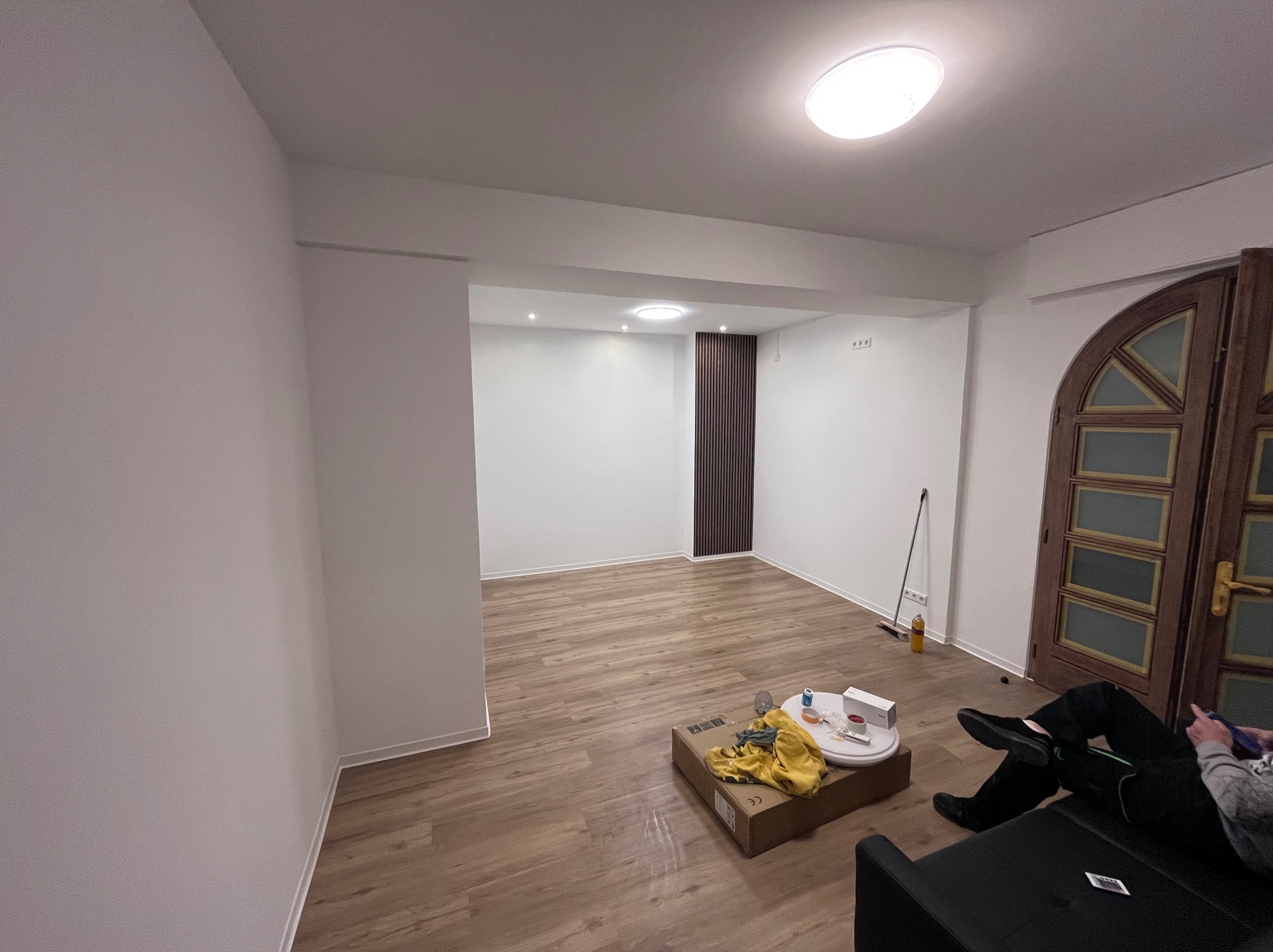Pokoje po remoncie z nową podłogą z paneli, widoczny fragment sofy z osobą, karton z narzędziami i akcesoriami malarskimi, oraz ozdobna ściana z pionowych listew.
