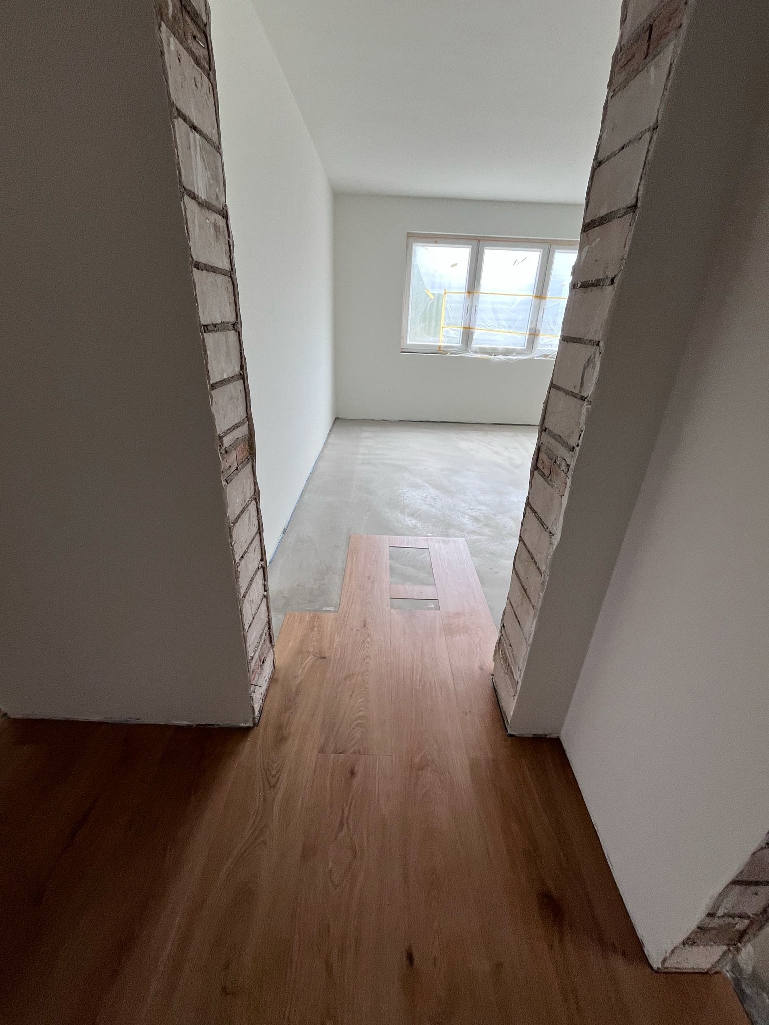 Fragment układania drewnopodobnych paneli; widoczne surowe wykończenie przejścia między pokojami i okno z folią ochronną w tle, w jasnym wnętrzu.