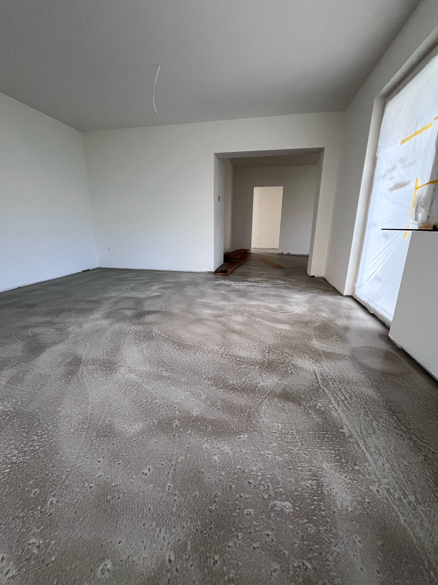 Wnętrze po wylaniu samopoziomującej wylewki, przygotowane do dalszych prac wykończeniowych. Okno zabezpieczone folią malarską, w tle widoczne kolejne pomieszczenie.