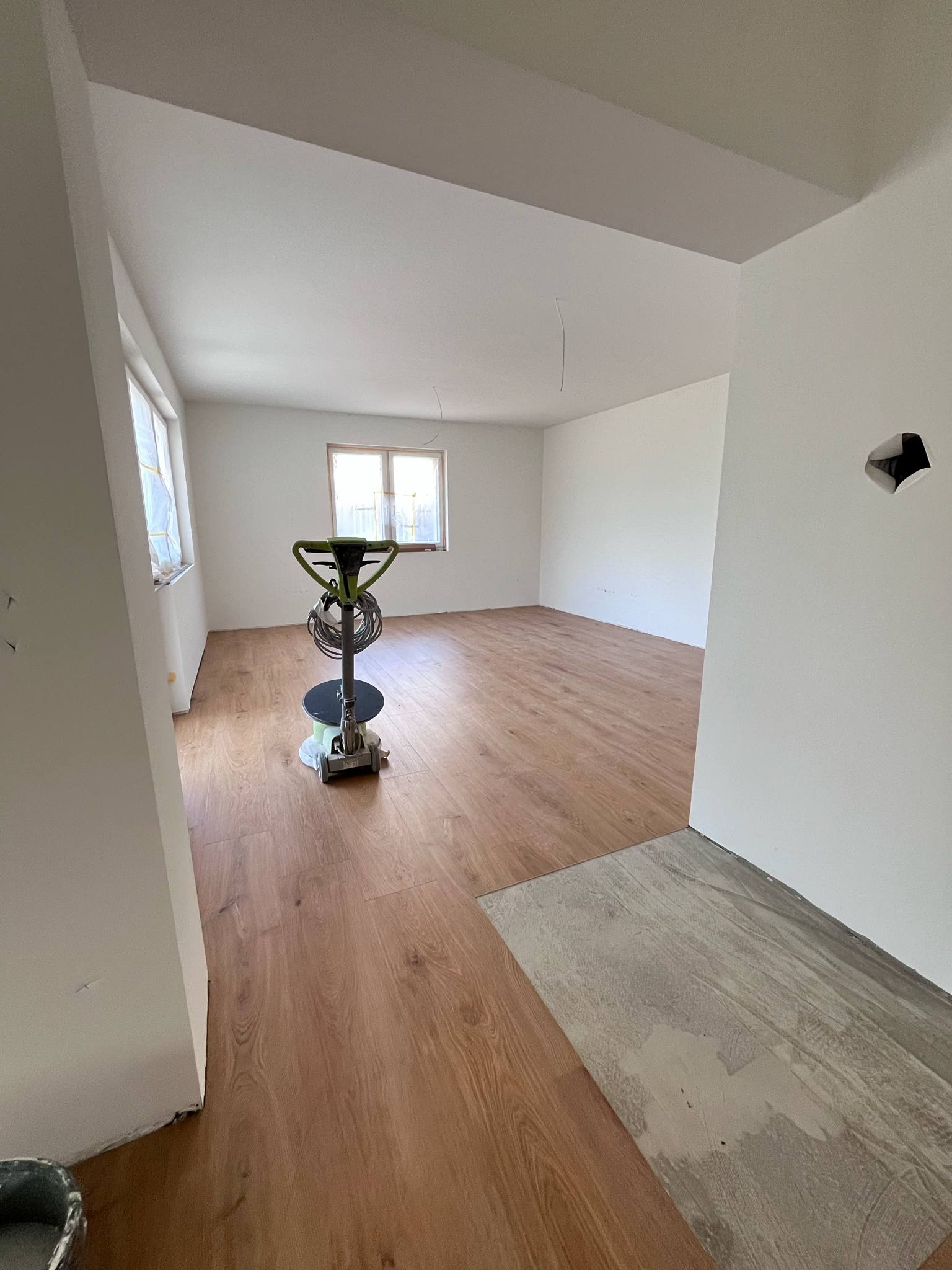 Parkiet w jasnym pokoju po cyklinowaniu, z maszyną do cyklinowania na pierwszym planie i fragmentem podłogi przed cyklinowaniem przy ścianie.