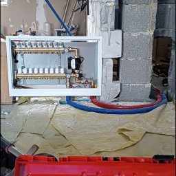 Konrad Gawęcki - Rozdzielacz ogrzewania podłogowego w białej obudowie, z niebieskimi i czerwonymi rurami. Widoczne narzędzia i czerwona skrzynka narzędziowa.