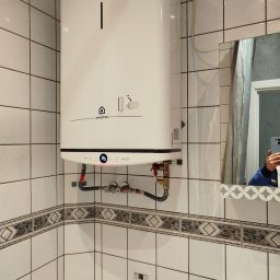 Konrad Gawęcki - Biały, wiszący bojler Ariston Velis w łazience z białymi płytkami i dekoracyjnym pasem. Widoczne przyłącza hydrauliczne i odbicie osoby w lustrze.