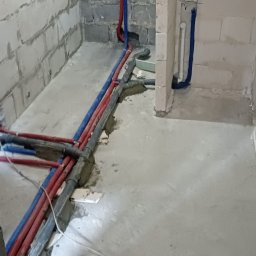 Konrad Gawęcki - Surowa łazienka w trakcie remontu z widocznymi rurami instalacji wodnej (czerwone i niebieskie) wychodzącymi ze ściany z betonu komórkowego.