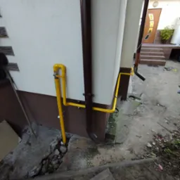 Żółta instalacja gazowa na elewacji budynku, z brązową rynną obok. Widoczne elementy mocowania i zawór. Fragment podwórka z betonową nawierzchnią.