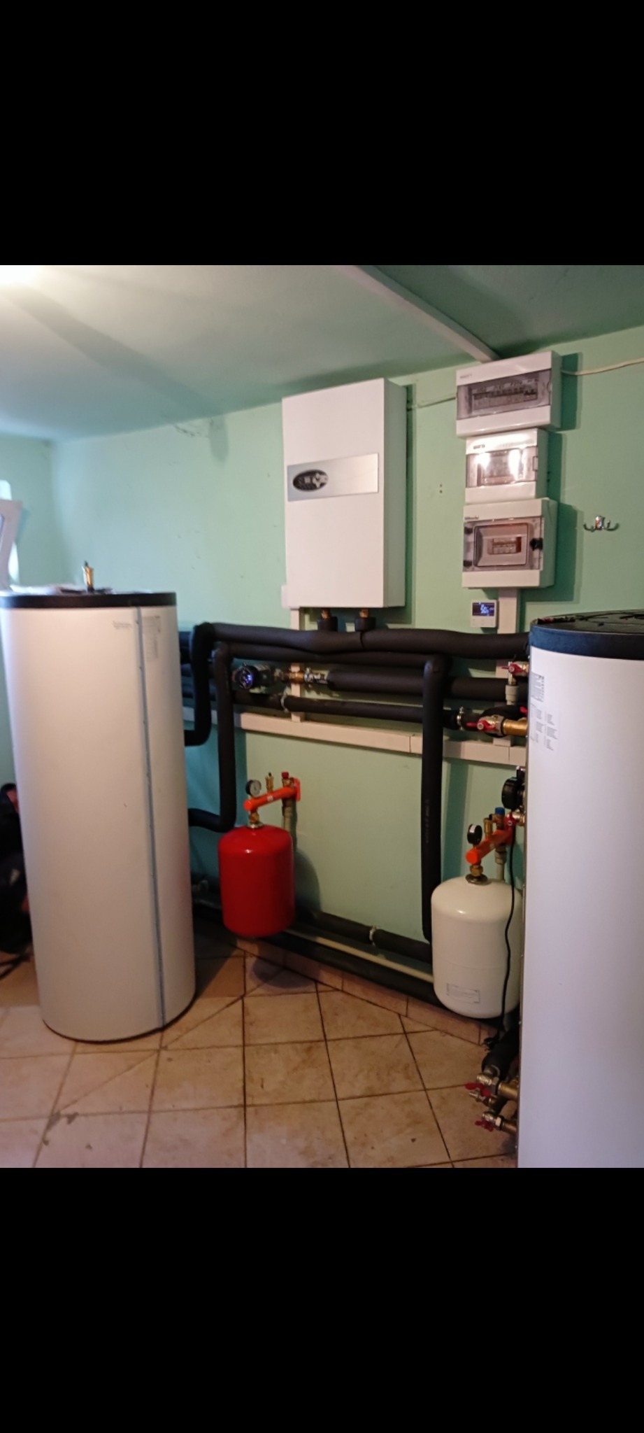 Instalacja CO z piecem, zbiornikami i rurami w piwnicy. Widoczne zawory, manometry i skrzynka elektryczna na ścianie. Całość w pomieszczeniu z płytkami na podłodze.