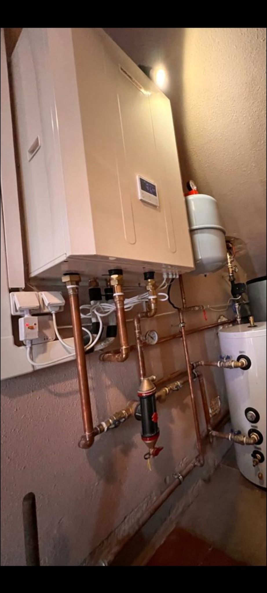 Biały piec CO z podłączeniem hydraulicznym, rurami miedzianymi i zbiornikiem wyrównawczym. Widoczny filtr i zawory. Całość zamontowana na ścianie.