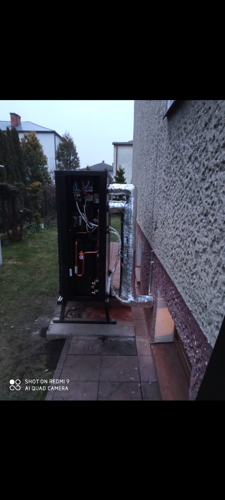 Zewnętrzna pompa ciepła zamontowana przy ścianie budynku, z widocznymi rurami w izolacji. Widok z boku, na tle trawnika i zabudowań.