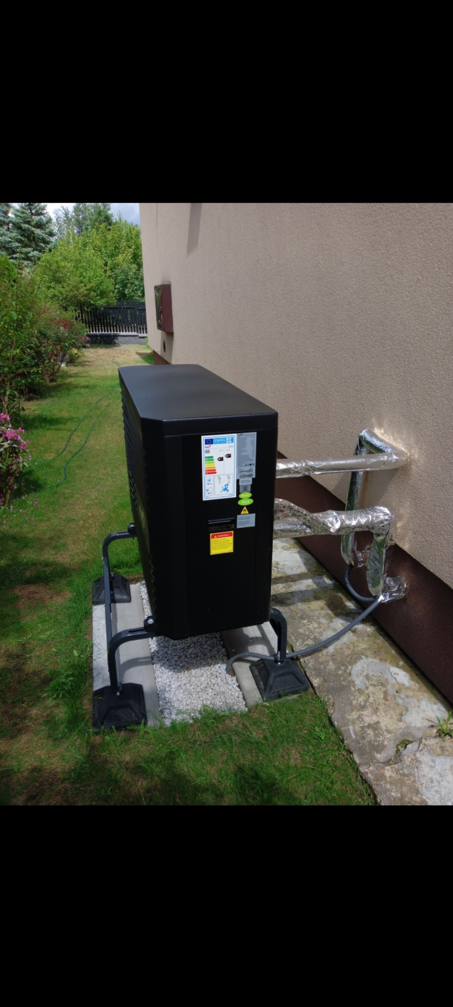Czarna pompa ciepła zamontowana na zewnątrz budynku z widocznymi rurami instalacji, na tle zielonego trawnika i ściany. Widoczne etykiety energetyczne na urządzeniu.