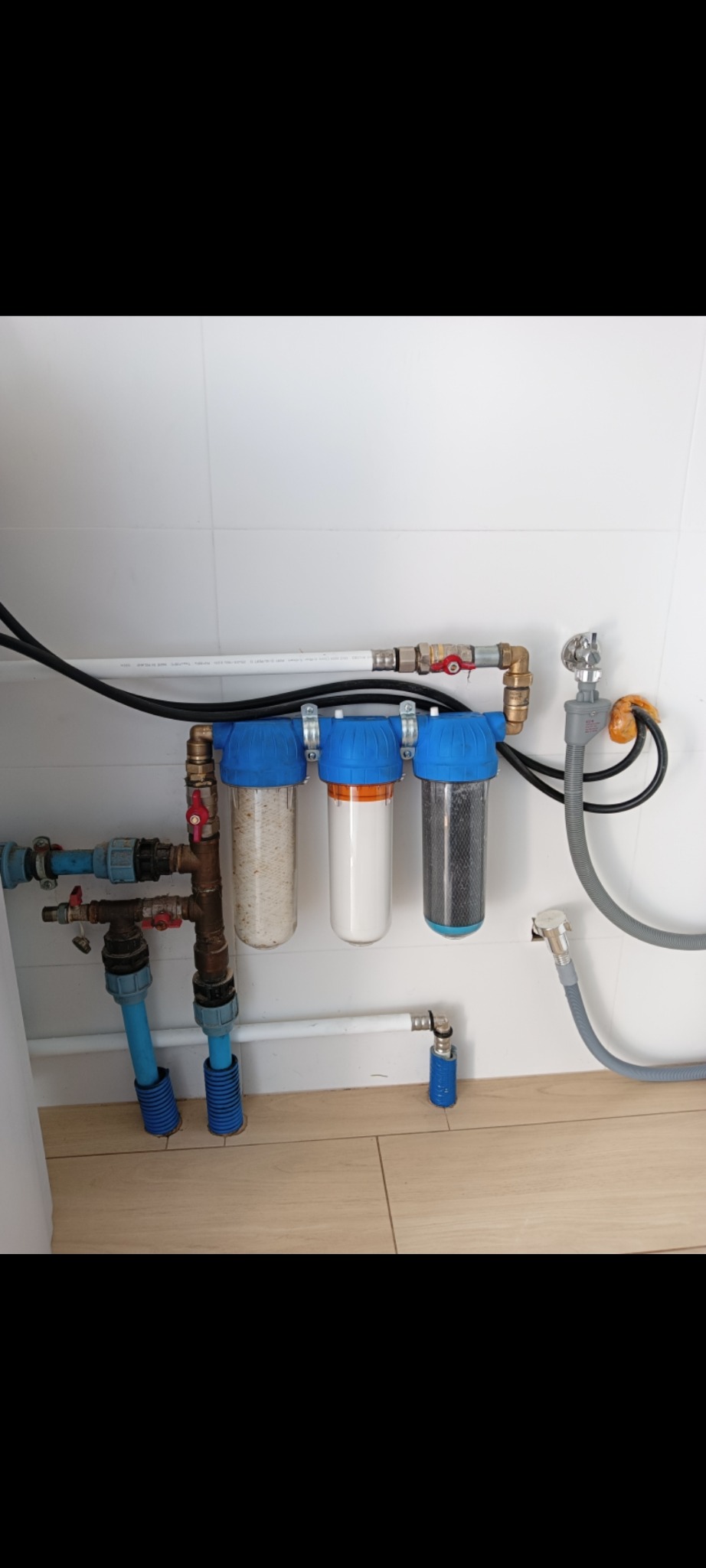 Zestaw filtrów wody z trzema przezroczystymi cylindrami i niebieskimi pokrywami zamontowany na ścianie nad podłogą z płytek, z widocznymi rurami i zaworami.