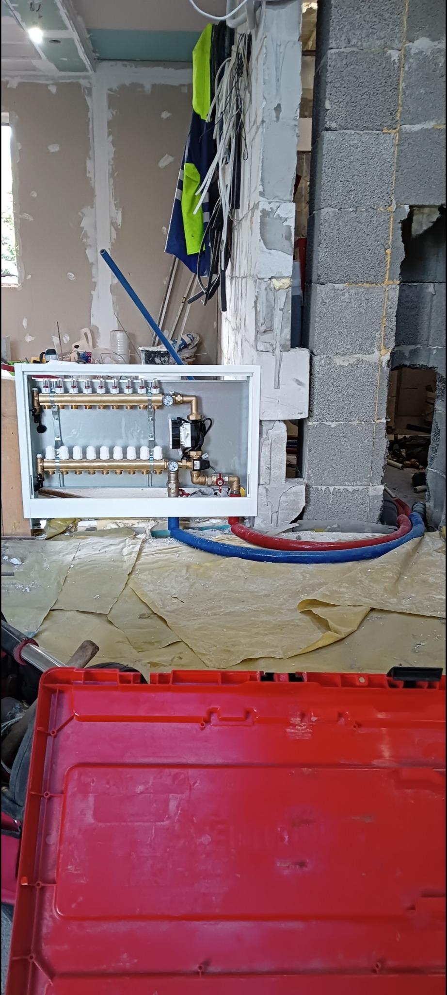 Rozdzielacz ogrzewania podłogowego w białej obudowie, z niebieskimi i czerwonymi rurami. Widoczne narzędzia i czerwona skrzynka narzędziowa.