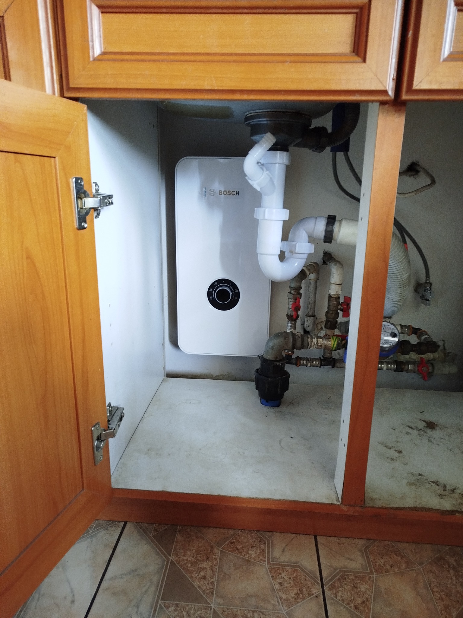 Biały piec Bosch w szafce pod zlewem, widoczne rury i zawory. Otwarta drewniana szafka z widocznymi zawiasami. Widok na podłogę z płytek.