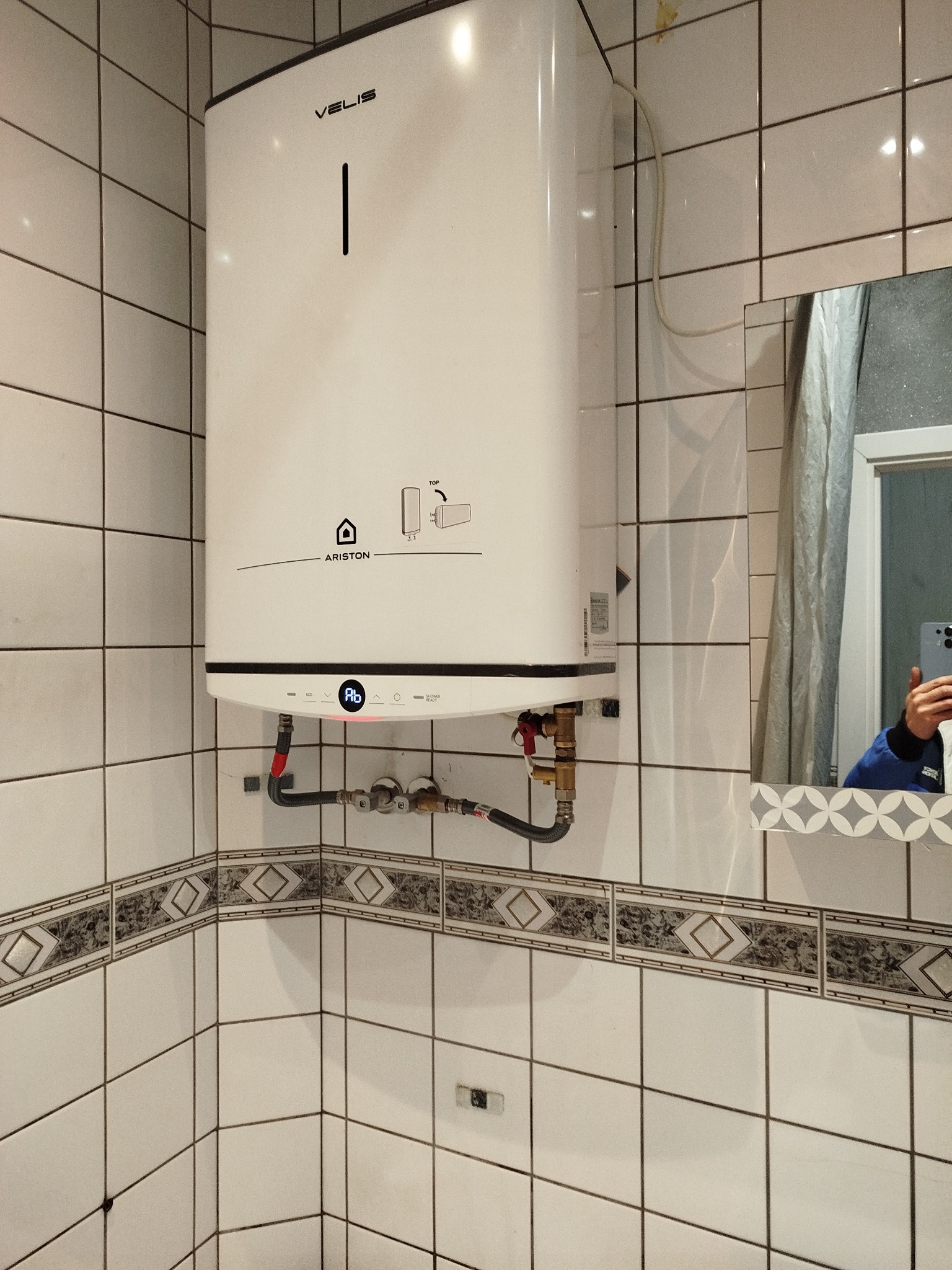 Biały, wiszący bojler Ariston Velis w łazience z białymi płytkami i dekoracyjnym pasem. Widoczne przyłącza hydrauliczne i odbicie osoby w lustrze.