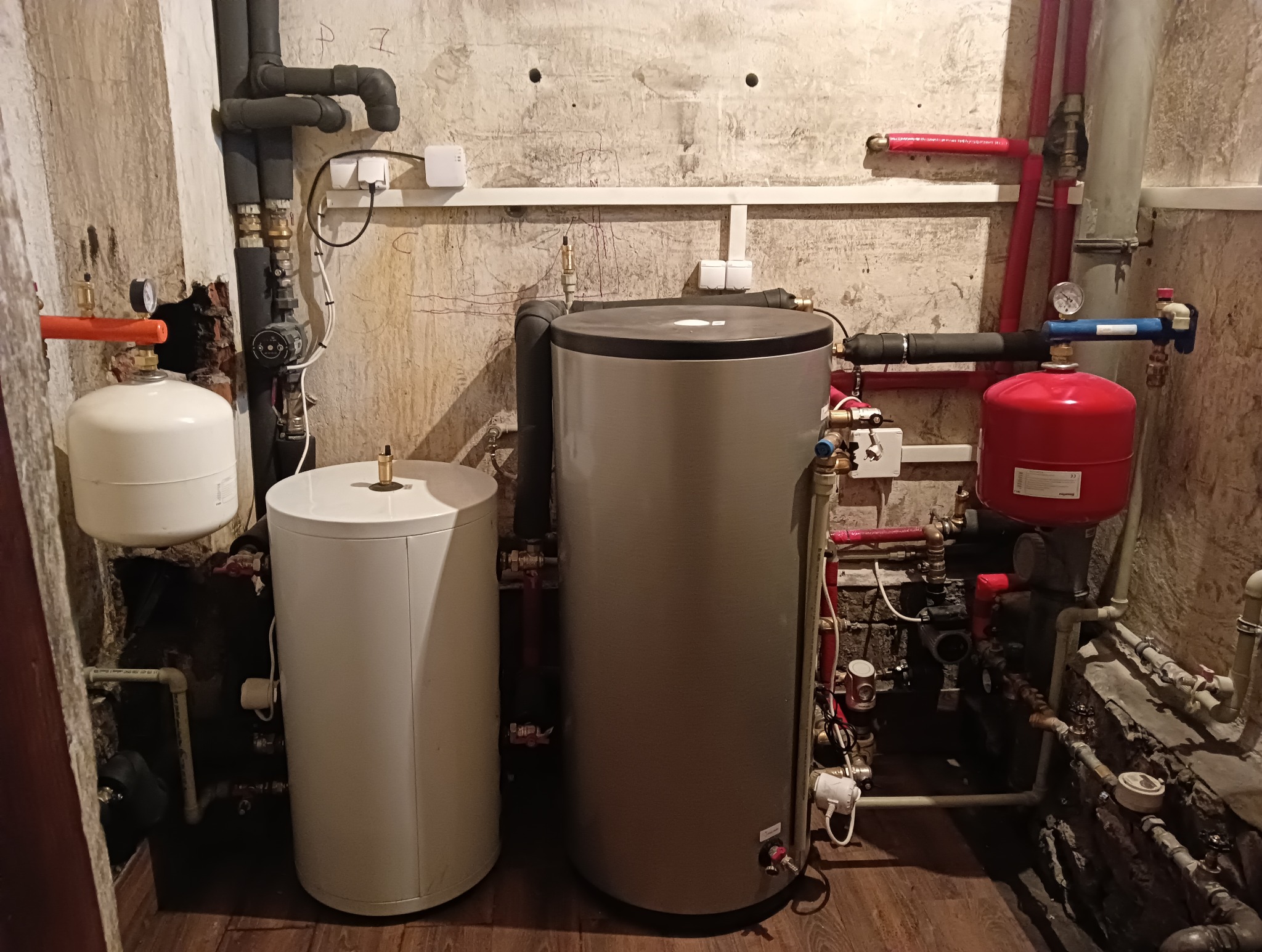 Dwa zbiorniki: biały i szary, obok czerwonego naczynia przeponowego. Widoczne rury, zawory i instalacja w pomieszczeniu o surowych ścianach.