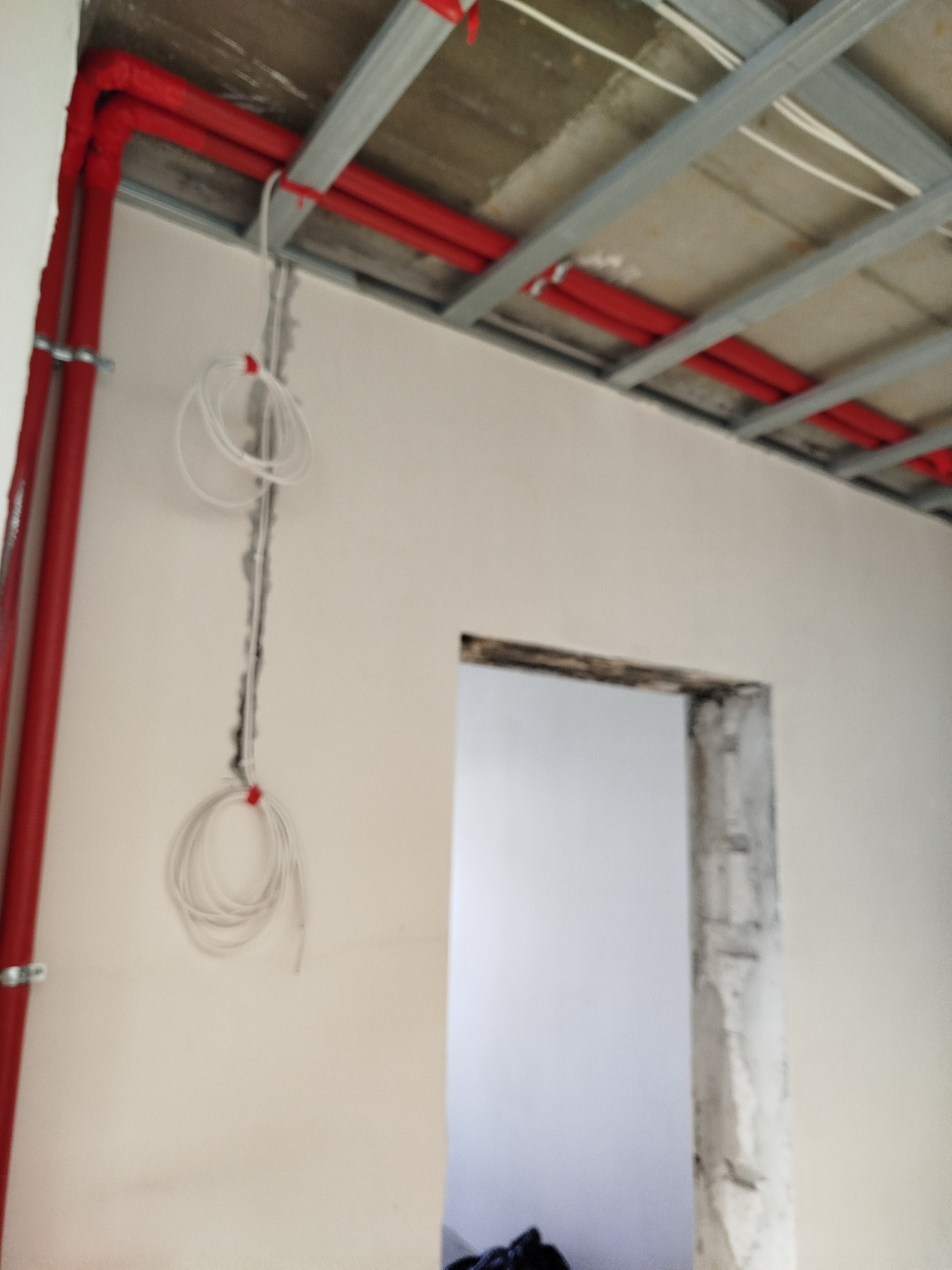 Czerwone rury instalacji pod stropem, białe przewody na ścianie, widoczne metalowe profile sufitowe i fragment ościeżnicy drzwiowej. Prace instalacyjne w trakcie.