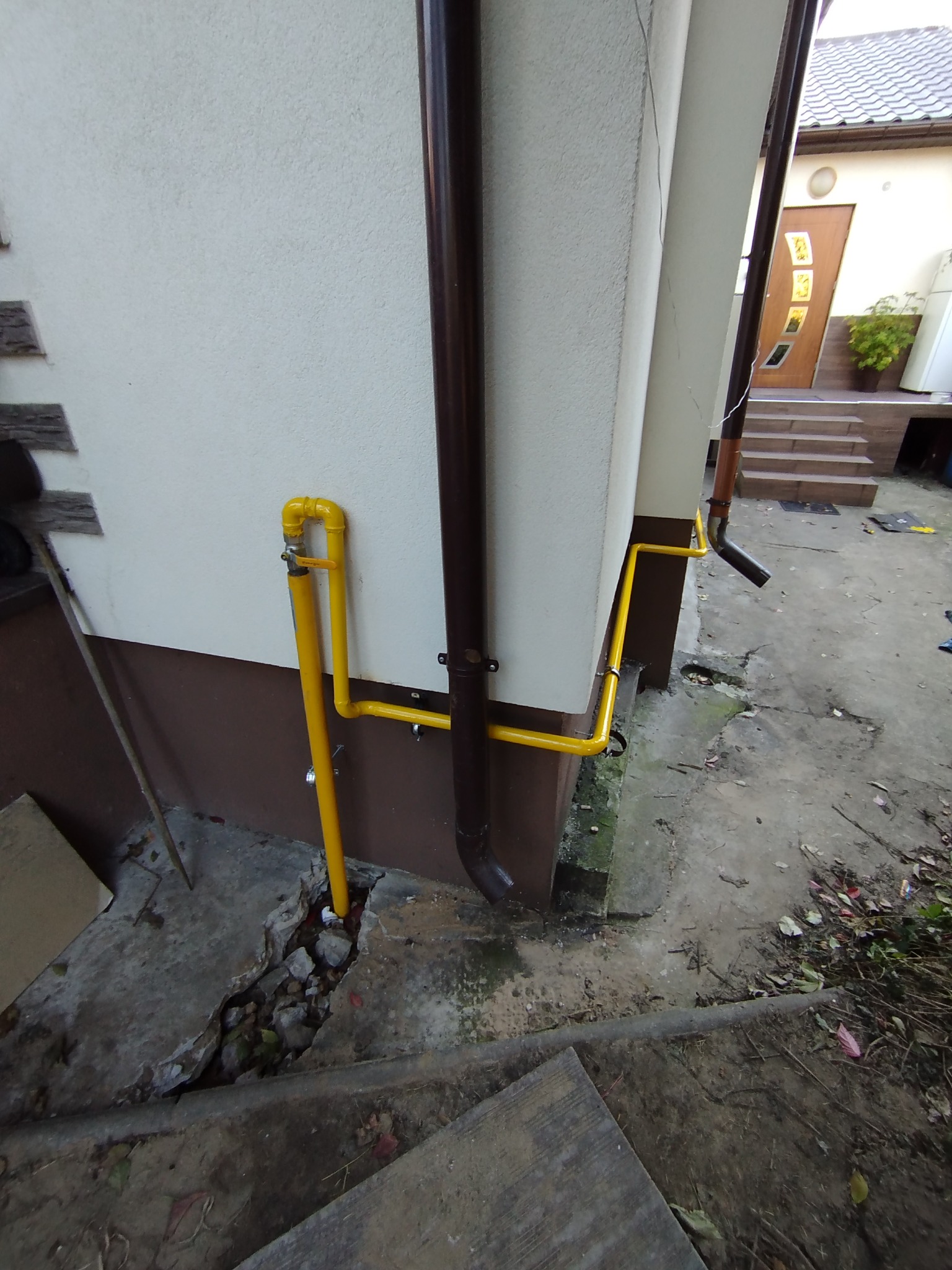 Żółta instalacja gazowa na elewacji budynku, z brązową rynną obok. Widoczne elementy mocowania i zawór. Fragment podwórka z betonową nawierzchnią.
