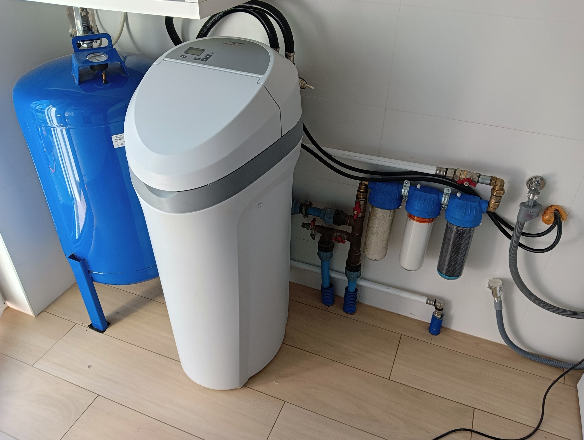 Zestaw filtrów wody z niebieskim zbiornikiem ciśnieniowym i białym zmiękczaczem, zainstalowany przy ścianie z białych płytek i podłodze imitującej drewno.