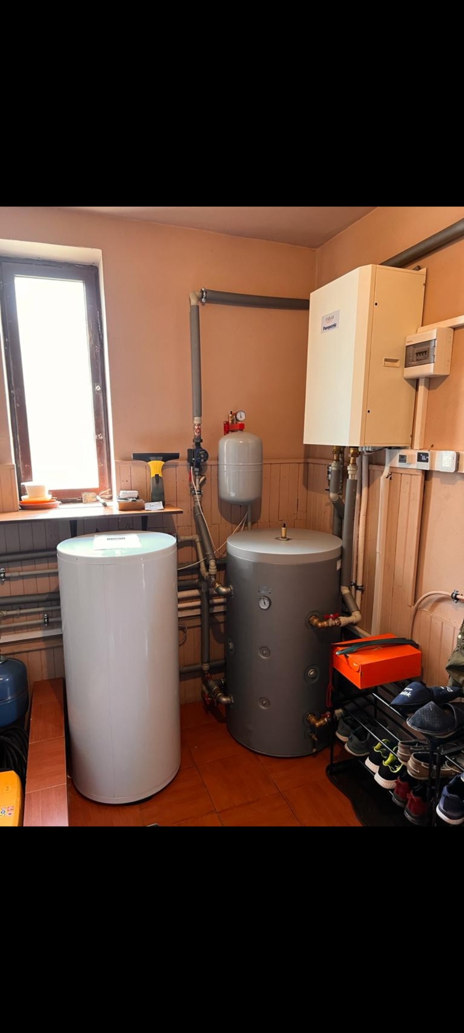 Biały i szary zasobnik ciepłej wody użytkowej w kotłowni, obok okna z akcesoriami. Na ścianie piec gazowy i instalacja. Na podłodze brązowe płytki.