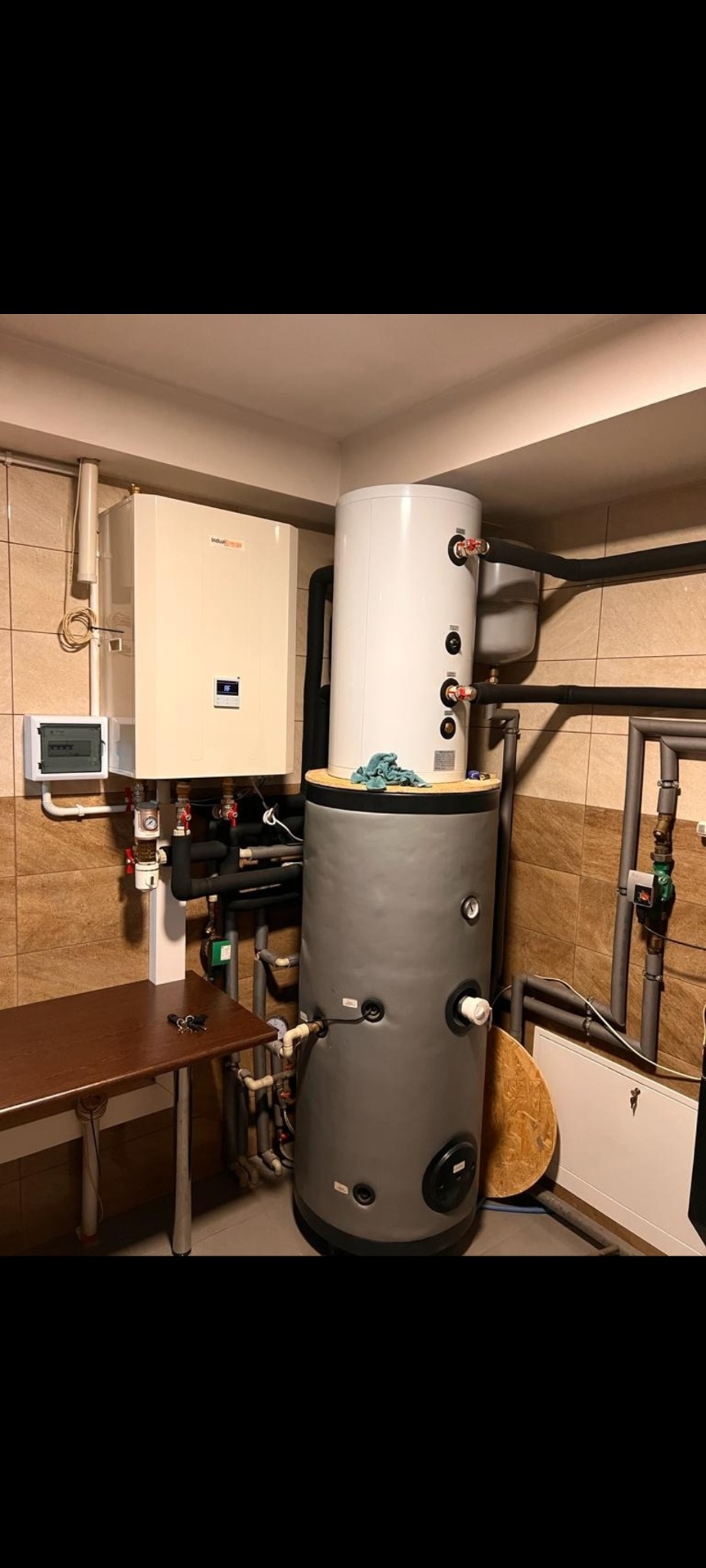 Biały piec centralnego ogrzewania i szary zbiornik na wodę w kotłowni z brązowymi płytkami. Widoczne rury, zawory i brązowy stolik.