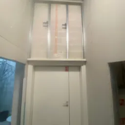 Wnętrze w trakcie wykończenia: białe ściany z widocznymi przewodami elektrycznymi nad drzwiami, metalowa konstrukcja pod zabudowę. Stan surowy, przygotowania do dalszych prac.