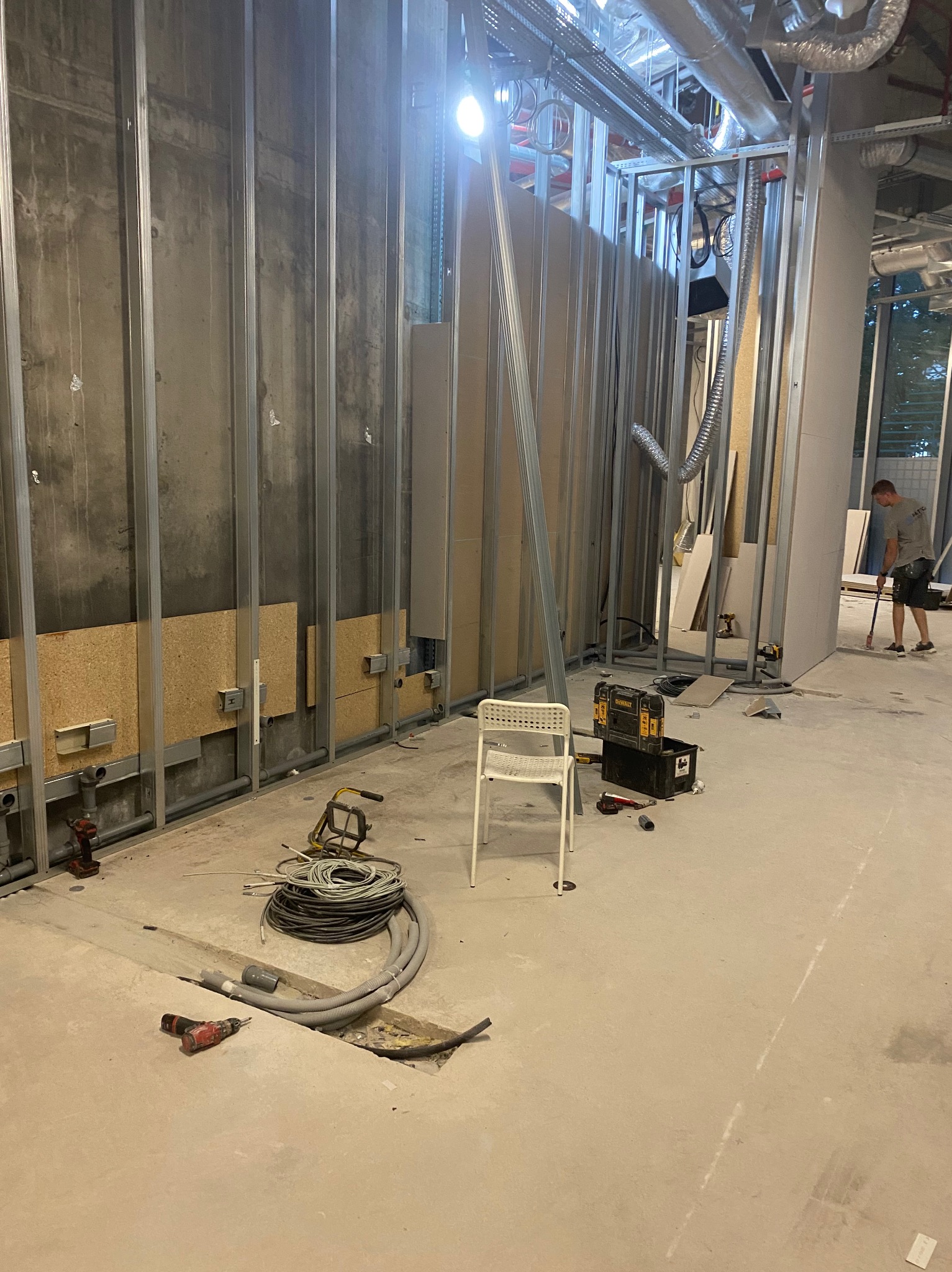 Wnętrze w trakcie prac wykończeniowych: metalowy szkielet ścian działowych, częściowo obłożony płytami, instalacje, narzędzia i pracownik w tle.