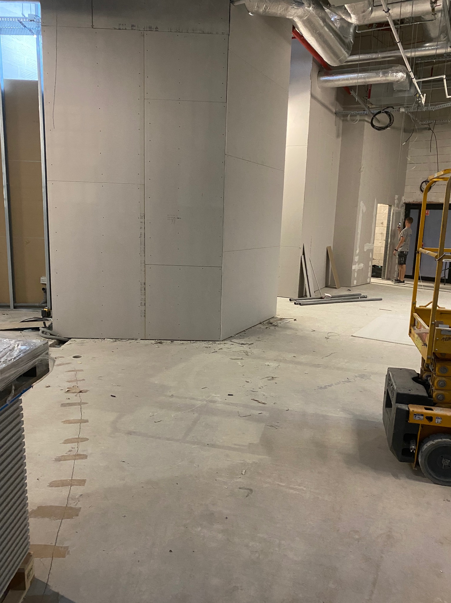 Montaż płyt gipsowo-kartonowych na ścianach w przestrzeni komercyjnej, widoczny podnośnik i elementy instalacji pod sufitem. Prace wykończeniowe w toku.