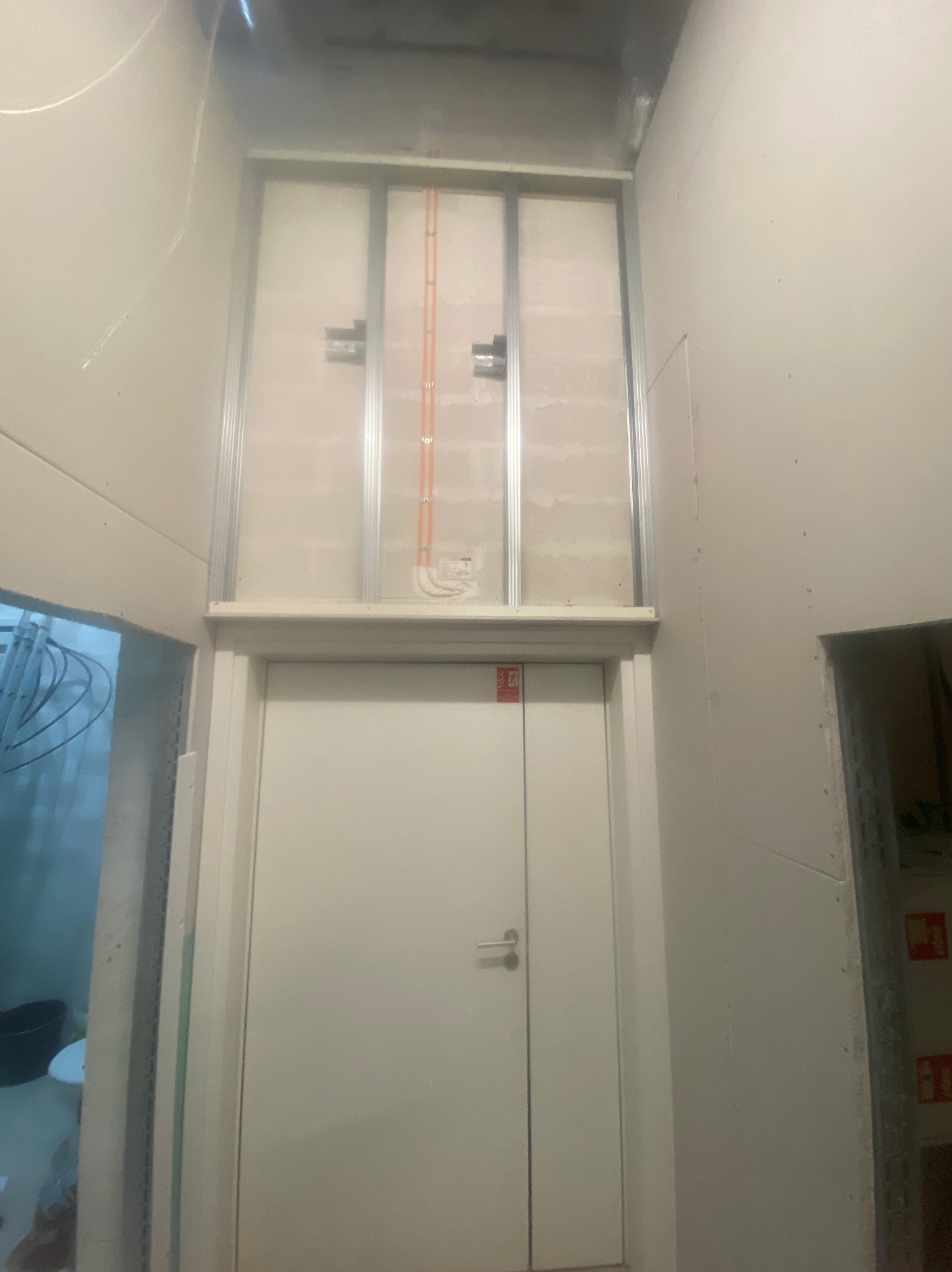 Wnętrze w trakcie wykończenia: białe ściany z widocznymi przewodami elektrycznymi nad drzwiami, metalowa konstrukcja pod zabudowę. Stan surowy, przygotowania do dalszych prac.