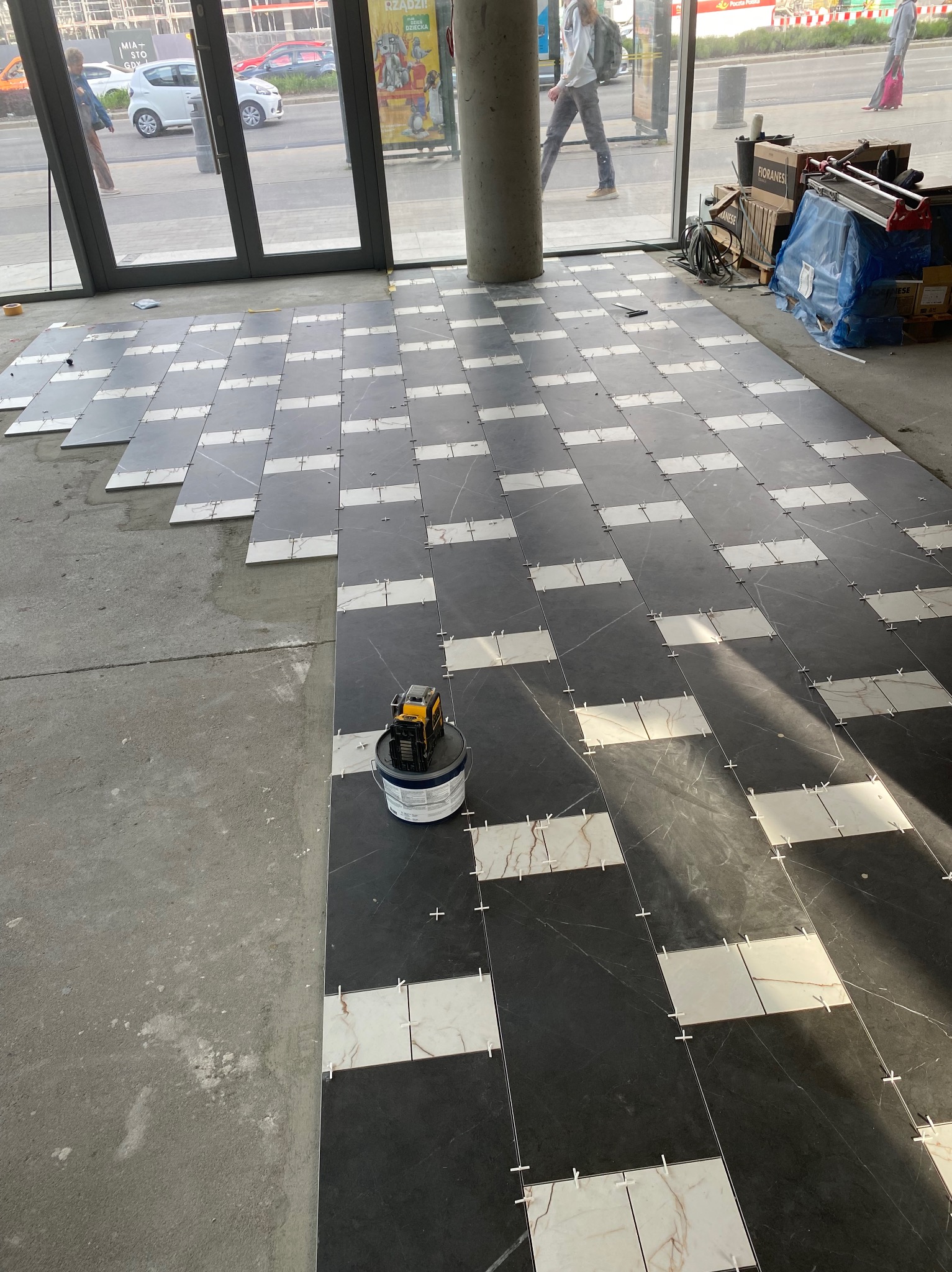 Układanie płytek podłogowych w szachownicę, z użyciem krzyżyków dystansowych i poziomicy laserowej, na surowym podłożu w jasnym wnętrzu.