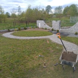 Szpad_bruk - Układanie ścieżki z kostki granitowej w ogrodzie; widoczny fragment muru z kamienia, brama z kutego żelaza, taczka z ziemią i pracownik z kielnią.