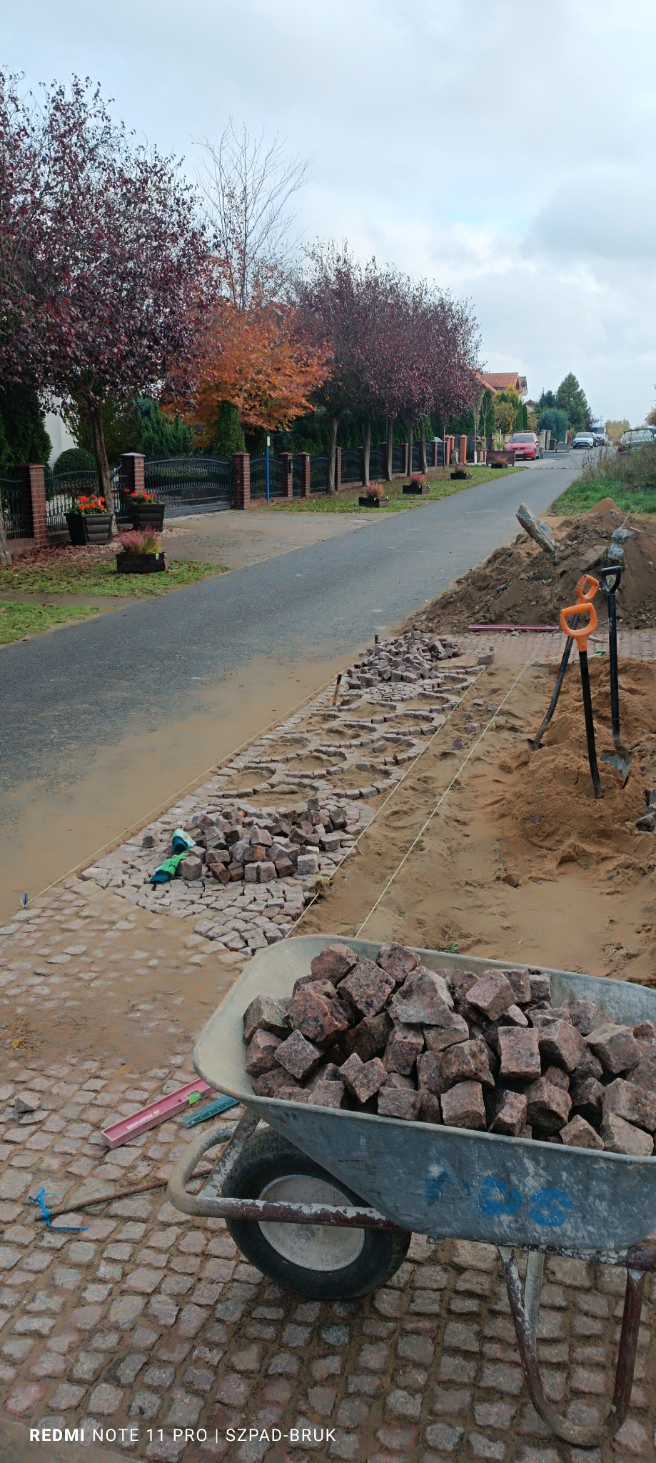 Ułożona częściowo nawierzchnia z kostki granitowej wzdłuż ulicy, taczka wypełniona kostką, przygotowane podłoże z piasku, widoczne narzędzia: łopaty, poziomica, sznurek wyznaczający linię. W tle...