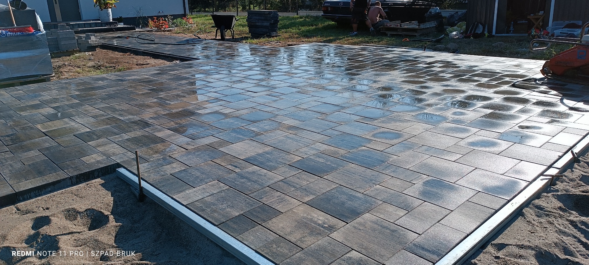 Układanie kwadratowej kostki brukowej w odcieniach szarości, mokra powierzchnia odbija światło, widoczna listwa krawężnikowa i ubijarka w tle.