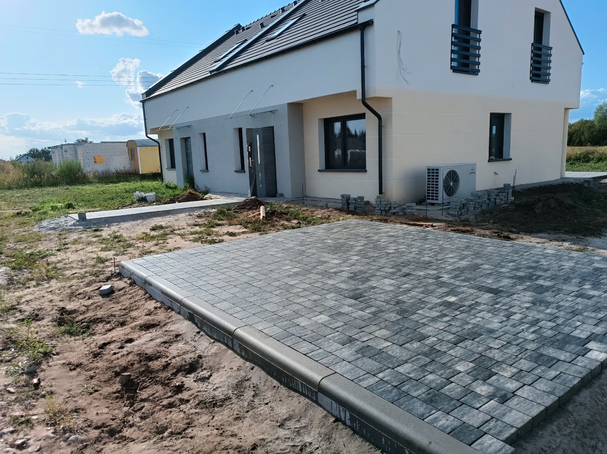 Częściowo ułożona, szara kostka brukowa z obrzeżem na posesji przed nowoczesnym, dwukondygnacyjnym domem z klimatyzatorem na ścianie bocznej i stertą kostki obok.