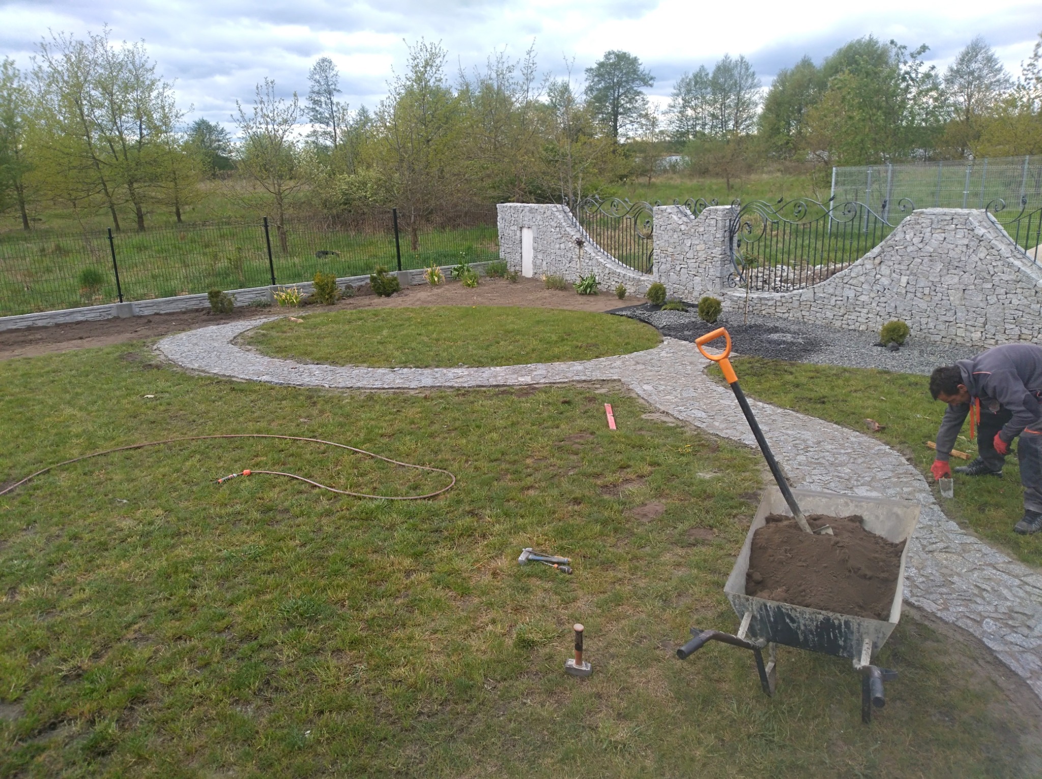 Układanie ścieżki z kostki granitowej w ogrodzie; widoczny fragment muru z kamienia, brama z kutego żelaza, taczka z ziemią i pracownik z kielnią.