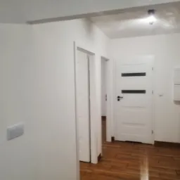 Wnętrze przedpokoju po remoncie: białe ściany z włącznikiem światła, drewnopodobna podłoga, trzy białe drzwi z czarnymi akcentami, oświetlenie punktowe.