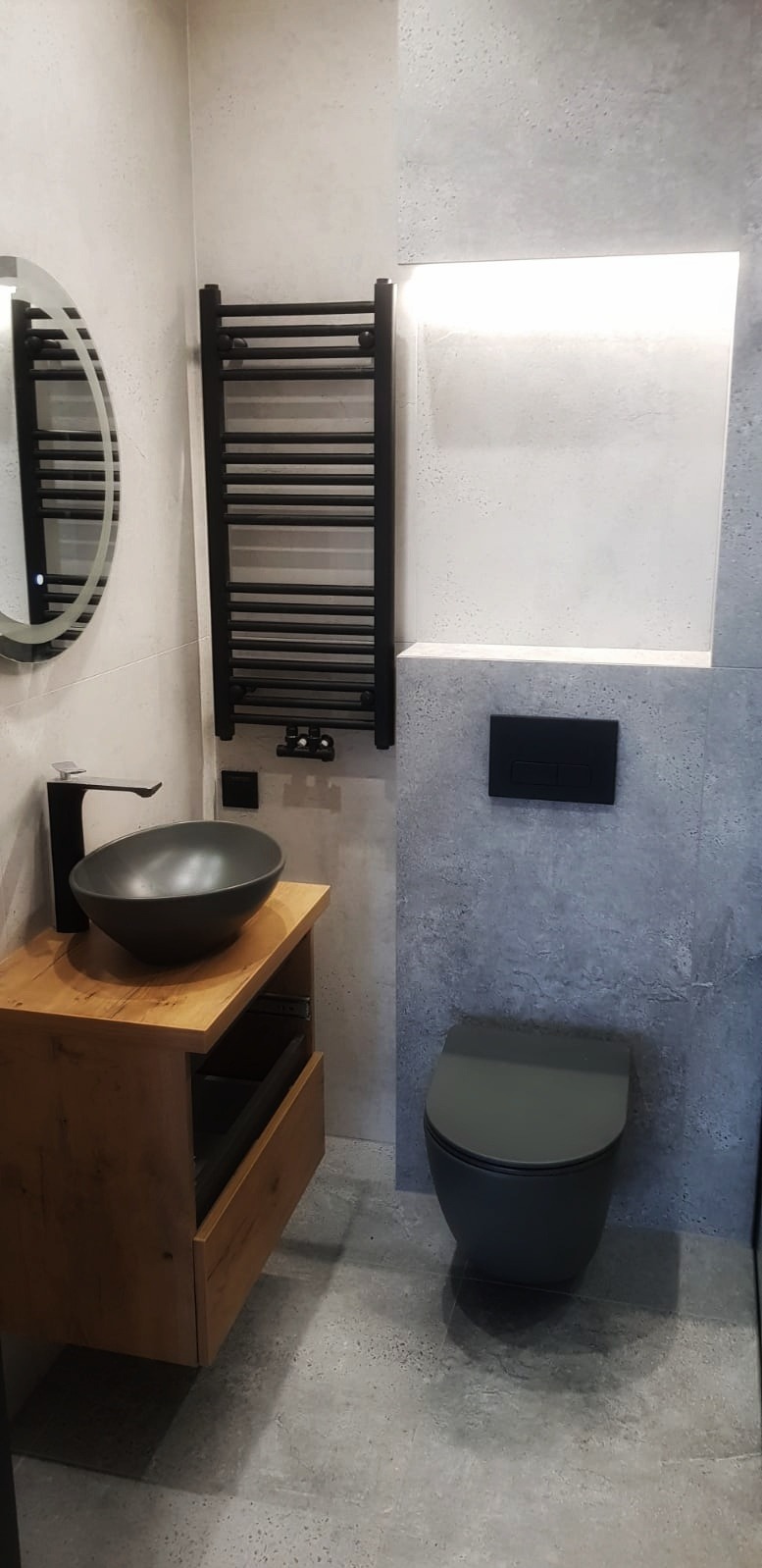 Nowoczesna łazienka z umywalką w kształcie misy na drewnianej szafce, czarnym grzejnikiem drabinkowym i podwieszaną toaletą w odcieniach szarości.